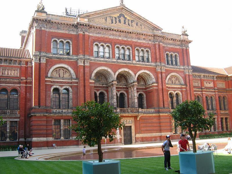The ornate red brick and terracotta facade of the Victoria and Albert Museum's courtyard in London, featuring arched windows and classical sculptures, overlooks a green lawn with a shallow pool and two potted trees.