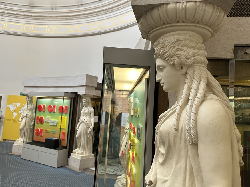 A side profile of a white caryatid statue in the foreground, with two similar statues framing a museum exhibit about cryptoassets in the background.