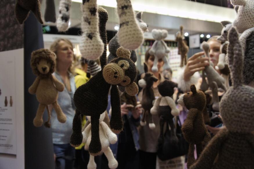 A variety of small, handcrafted crochet animals, including a monkey, lion, and giraffe, hang from the ceiling at a busy indoor market.