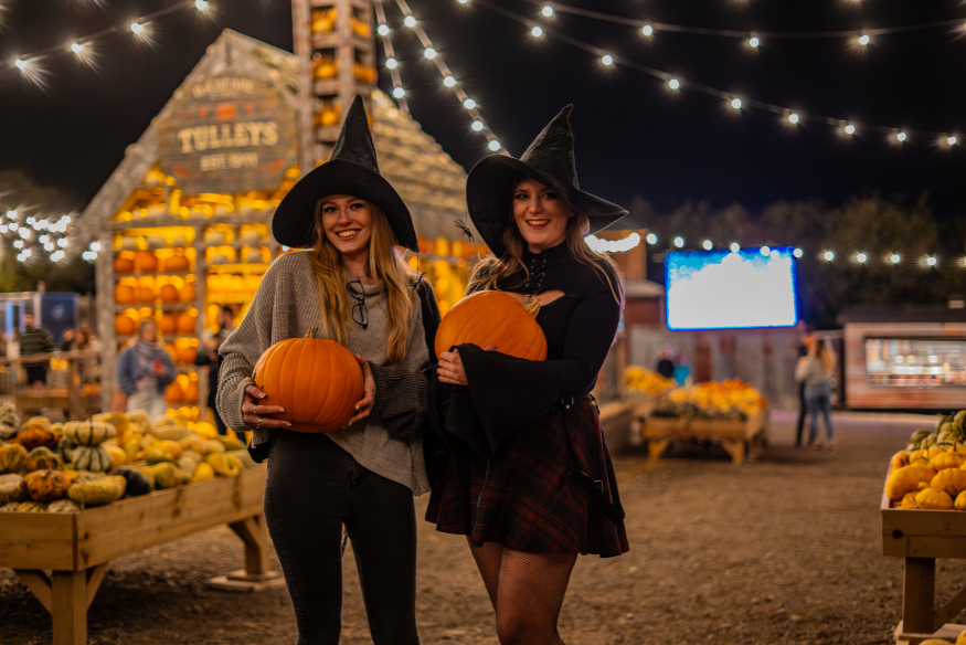 Pumpkin picking farms and patches near London 2026: two women dressed as witches holding pumpkins in a field lit by fairy lights at night