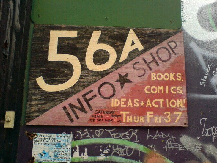 A weathered wooden sign for 56a Infoshop, a radical social center, featuring hand-painted text and a diagonal split between black and red backgrounds. The sign lists "Books, Comics, Ideas + Action!" along with opening hours, set against a wall covered in graffiti.