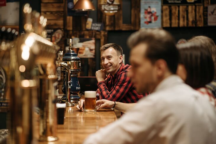 A man in a red plaid shirt smiles while sitting at a wooden bar counter with a glass of beer, as other patrons sit blurred in the foreground.
