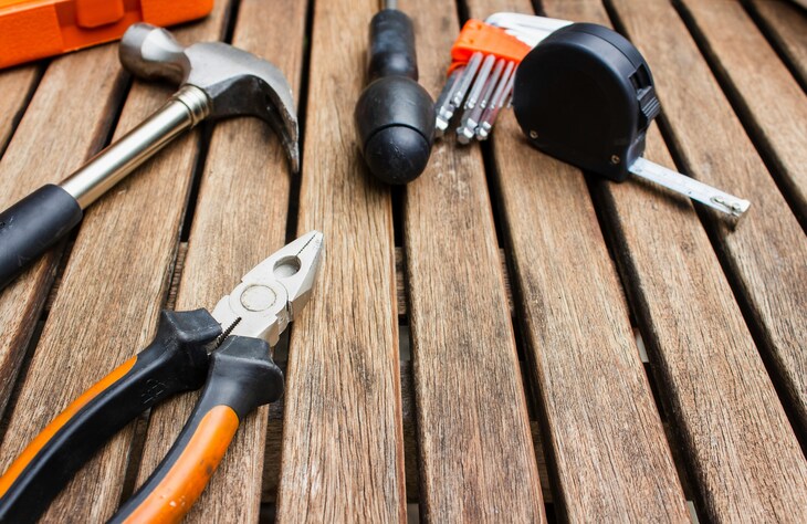 A collection of hand tools, including a hammer, pliers, a screwdriver, a set of hex keys, and a tape measure, scattered on a slatted wooden surface.