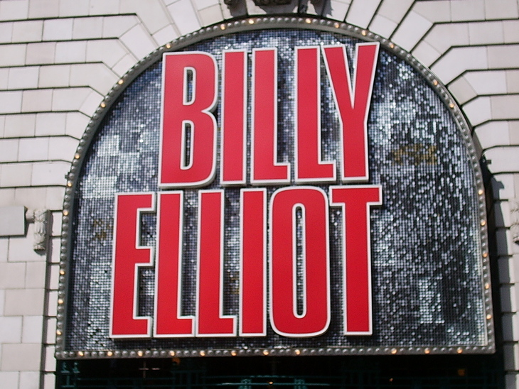 A large, arched theater sign for the musical "Billy Elliot" features bold red letters with white outlines against a shimmering silver sequined background.