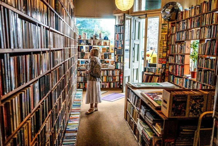 A woman with a backpack stands in a narrow aisle of a cozy, crowded bookstore, surrounded by floor-to-ceiling shelves packed with books and comics. Sunlight streams through an open doorway in the background.