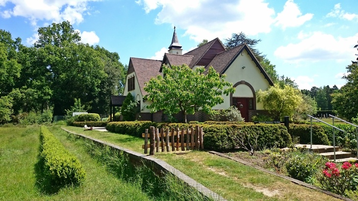 A small, cream-colored chapel with a brown shingled roof and a spire, surrounded by manicured hedges, green lawns, and lush trees under a bright blue sky.
