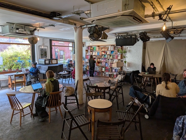 A dimly lit, industrial-style cafe and bookstore with exposed ceiling pipes and stage lights. People sit at scattered wooden tables working on laptops, while one person browses a large bookshelf in the background. A large window on the left looks out onto greenery.