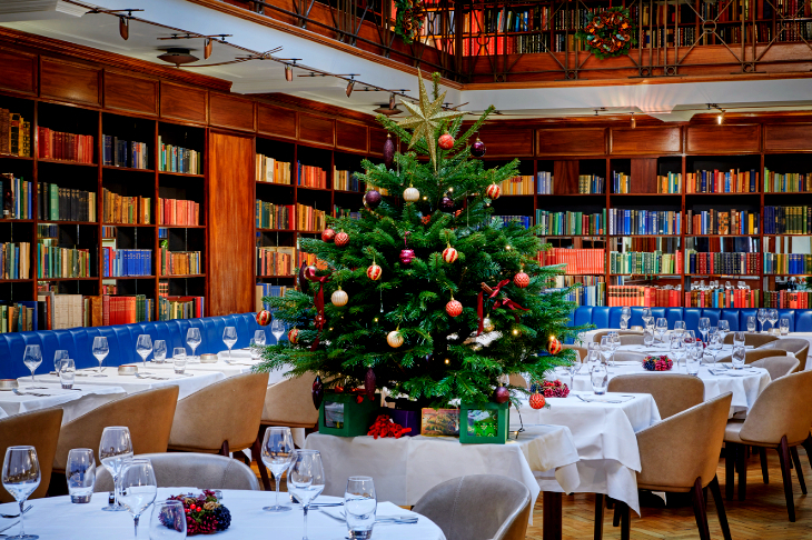Where to eat in London on Christmas Day 2026: a decorated Christmas tree on a table in the middle of the dining room. The wall around the edge of the dining room is lined with bookshelves.