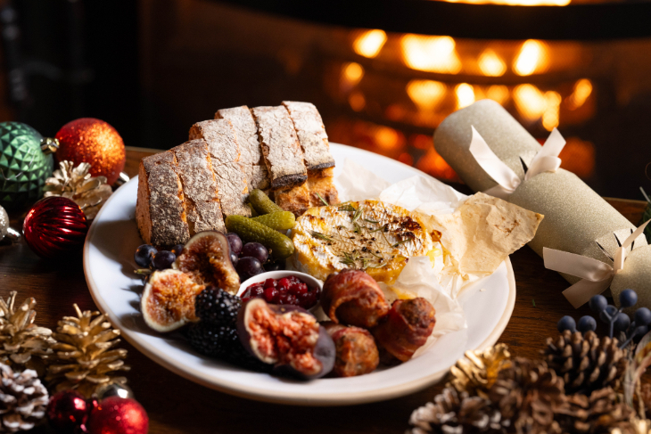 Where to eat in London on Christmas Day 2026: a table in front of a fireplace, decorated with pine cones and festive greenery, with a plate containing brie, cornichons, bread and figs