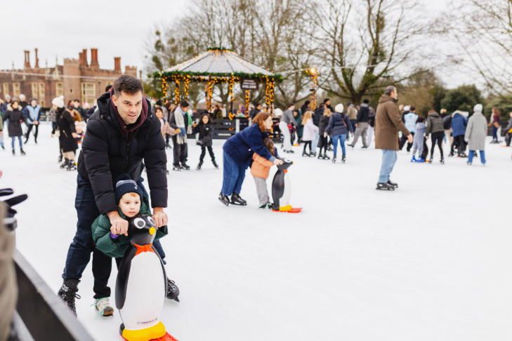 Things to do with kids in London at Christmas 2026: a young boy and a man ice skating together at Hampton Court Palace, holding on to a penguin skate aid