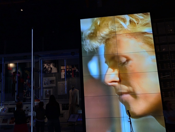 A large video screen in a dark museum exhibit displays a close-up profile of David Bowie with blonde hair, while visitors stand in the foreground looking at other displays.