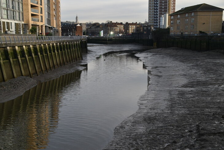 A narrow urban waterway at low tide, revealing muddy banks and a concrete retaining wall, with apartment buildings and a tall tower block in the background under an overcast sky.