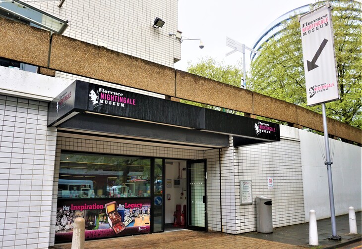 The entrance to the Florence Nightingale Museum, featuring a black awning with pink and white lettering, a large window display, and a tall banner sign with a downward arrow.