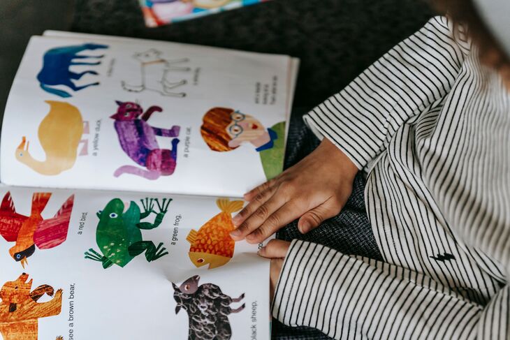 A child in a striped shirt looks at an open picture book featuring colorful illustrations of animals, including a purple cat, a green frog, and a brown bear.