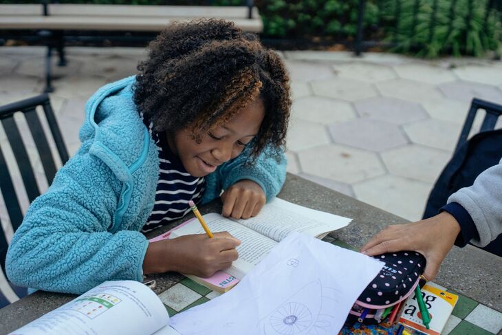 A young girl with curly hair and a blue fleece jacket smiles while writing in a book at an outdoor table.