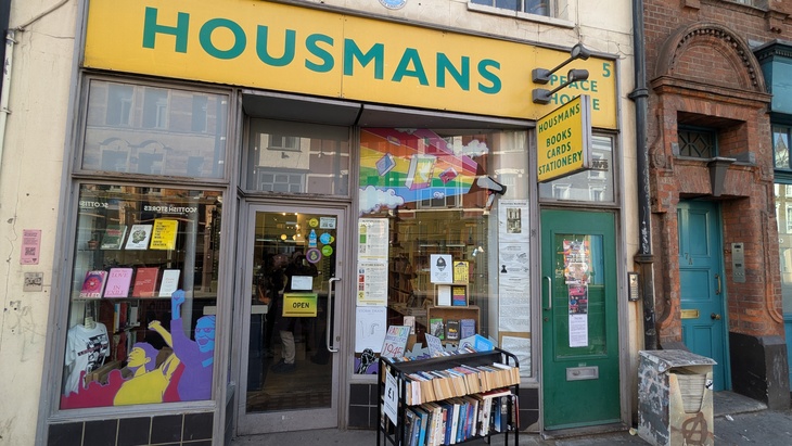 The storefront of Housmans Bookshop, featuring a large yellow sign with teal lettering, a window display with radical literature and rainbow graphics, and a green door to the right. A metal rack of books sits on the sidewalk in front of the window.