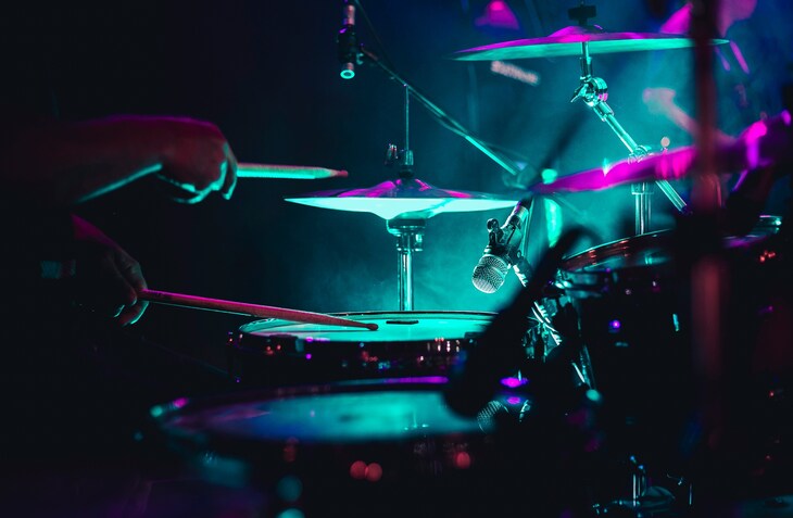 A close-up shot of a drummer playing a drum kit on a dark stage illuminated by vibrant teal and magenta stage lights.