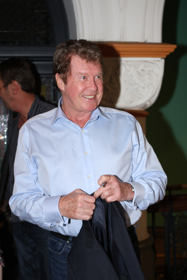 A smiling man with reddish-brown hair wearing a light blue button-down shirt and jeans, holding a dark jacket and a pen.