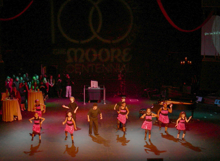 A group of young dancers in pink and black outfits perform on a stage at The Moore Theatre Centennial, with an audience and a piano in the background.