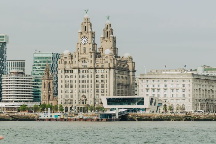 A wide shot of the Liverpool waterfront featuring the historic Royal Liver Building with its two clock towers and Liver Bird statues, flanked by modern skyscrapers and the white facade of the Cunard Building, all situated along the Mersey River.