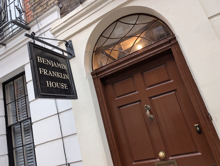 A low-angle shot of the entrance to Benjamin Franklin House in London, featuring a brown wooden door with a brass knocker and a black hanging sign with gold lettering.