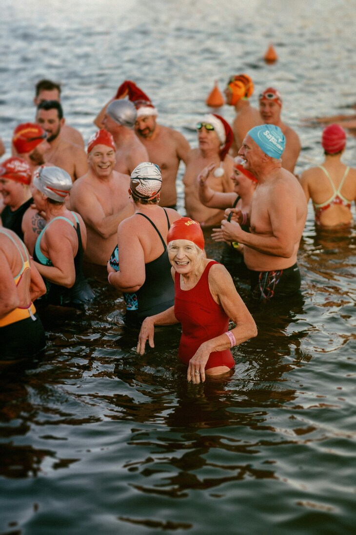 Things to do in London on Christmas Day 2026: A group of people, many wearing swim caps and some in Santa hats, smile and talk while wading in a body of water. In the foreground, an elderly woman in a red swimsuit and matching swim cap smiles directly at the camera.