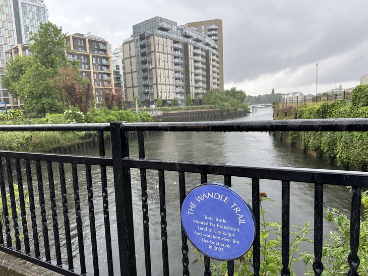 A view of the River Wandle in London on a rainy day, seen through a black metal railing with a blue circular plaque that reads, 