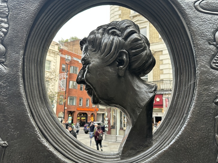 A bronze bust of Agatha Christie in profile, framed by a circular opening in a larger dark metal sculpture. The background shows a London street scene with pedestrians and buildings.