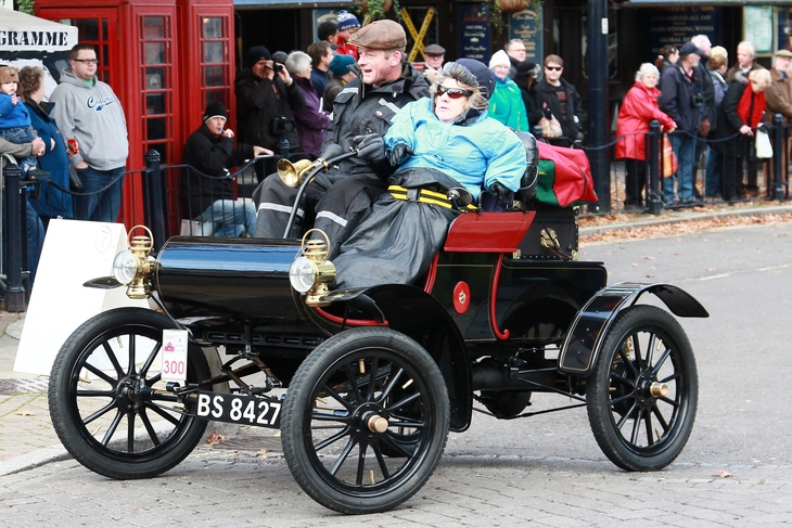 Things to do in London this weekend: A man and woman in period-style clothing drive a vintage black and red horseless carriage through a street lined with spectators.