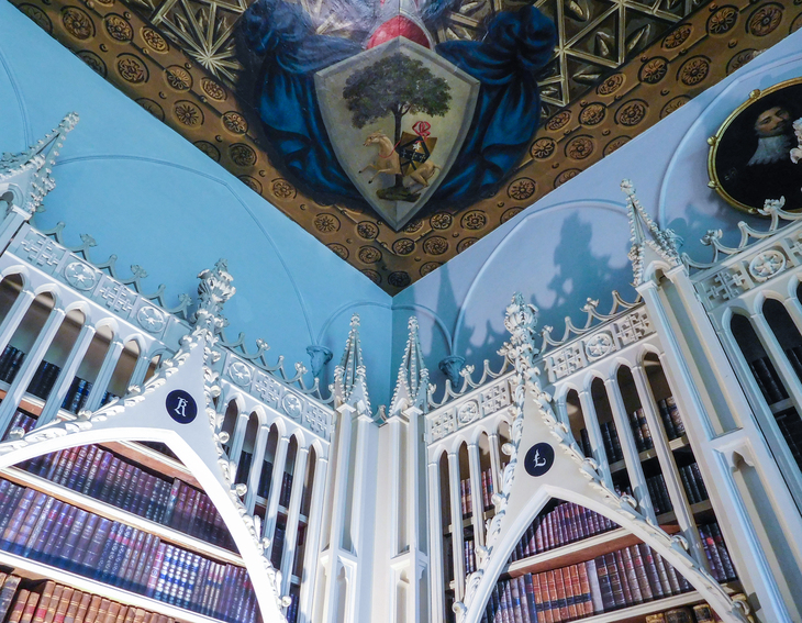 Things to do in London this weekend:  A low-angle shot of a library corner featuring white, Gothic-style arched bookshelves filled with leather-bound books. Above the shelves, a light blue wall meets a gold-patterned ceiling adorned with a large, painted coat of arms.
