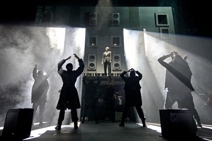 Things to do in London today: a photo of performers raising their arms in sync on stage, looking up at another person on a pedestal, with a backdrop of large speakers
