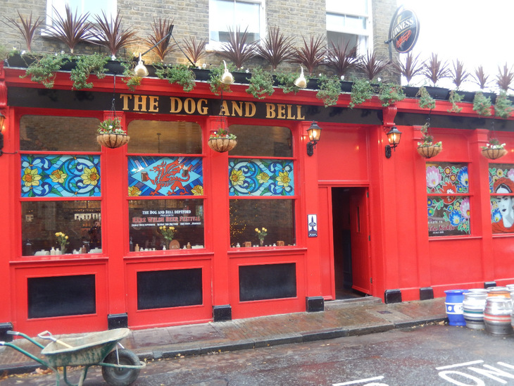 The bright red exterior of The Dog and Bell pub in Deptford, featuring stained glass windows, hanging flower baskets, and a wheelbarrow in front.