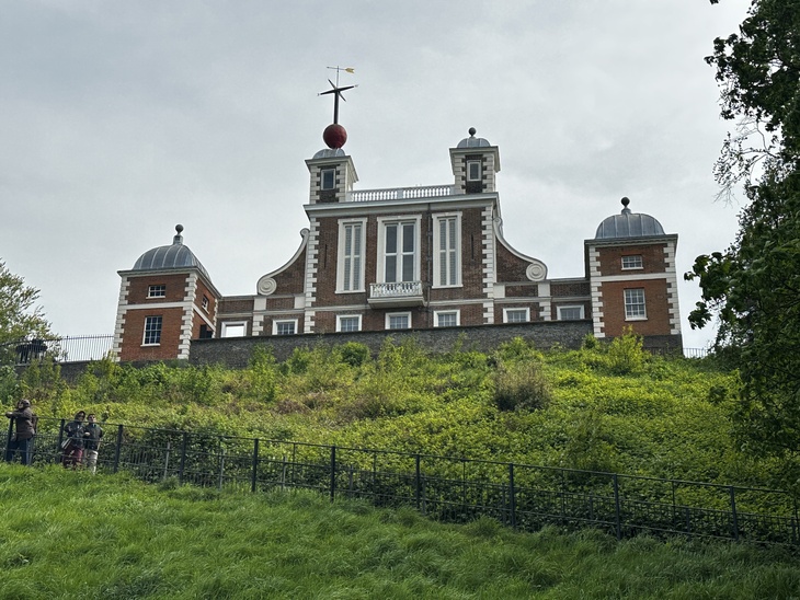 A low-angle shot of the Royal Observatory in Greenwich, London, featuring a red brick building with white trim and a red time ball on its roof, situated atop a grassy hill.