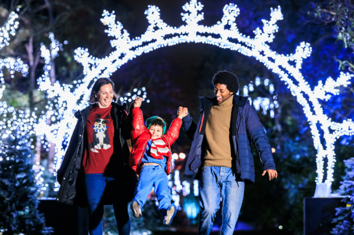 Things to do with kids in London at Christmas 2026: a man and woman swinging a child between them as they wander through an illuminated arch