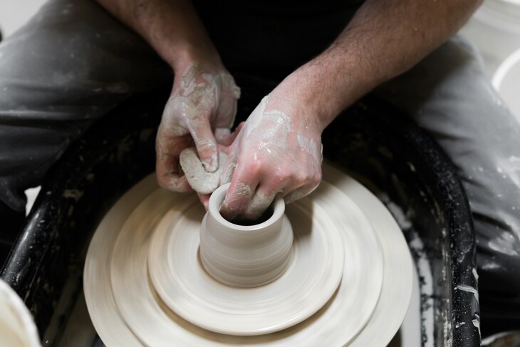 Close-up of a potter's clay-covered hands shaping a small ceramic vessel on a spinning pottery wheel.