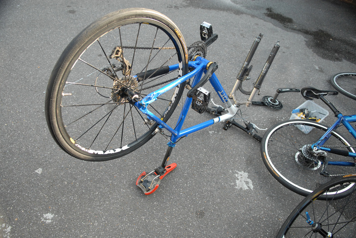 A blue and silver bicycle frame rests upside down on its seat and handlebars on an asphalt surface, with its front wheel removed and several other bike parts scattered nearby.