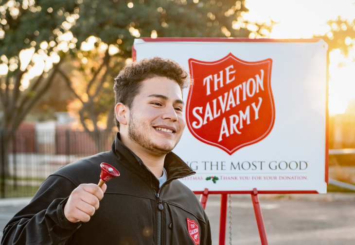 Volunteering in London, Christmas 2026: A smiling man in a black jacket stands next to a Salvation Army red kettle and sign, holding a small red bell.