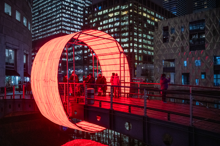 Canary Wharf Winter Lights 2027: people slihouetted walking through a glowing red ring on a footbridge over water
