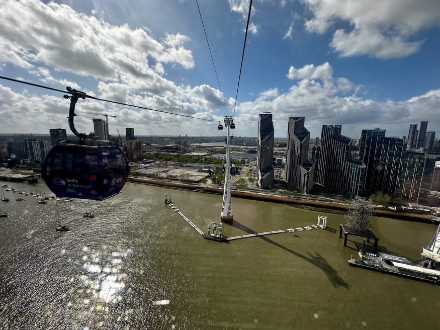 A view from a cable car over the River Thames in London, showing another purple gondola on the wire, a white support tower in the water, and modern skyscrapers on the riverbank under a bright, cloudy sky.