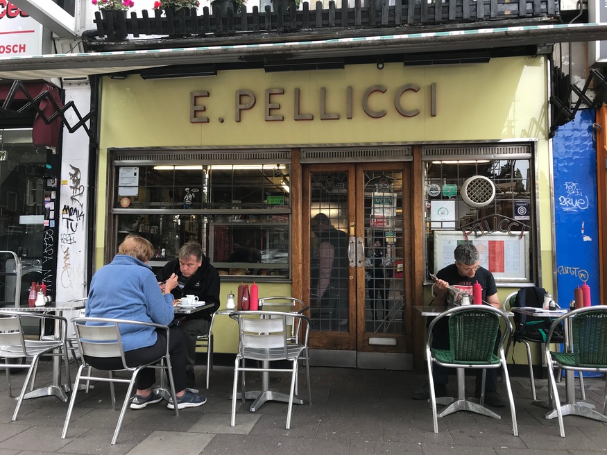 Three people sit at outdoor tables in front of E. Pellicci, a cafe with a yellow facade and wooden doors.