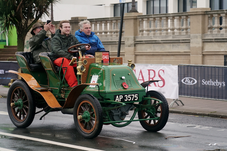 Three men ride in a vintage 1900 green Napier automobile with wooden wheels and brass accents during a street parade.