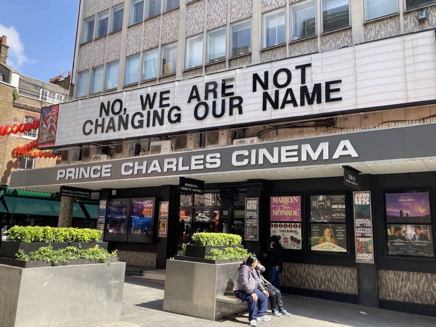 The Prince Charles Cinema in London with a marquee that reads, "NO, WE ARE NOT CHANGING OUR NAME," a humorous reference to the accession of King Charles III. Movie posters and people sitting on a bench are visible in front of the building.