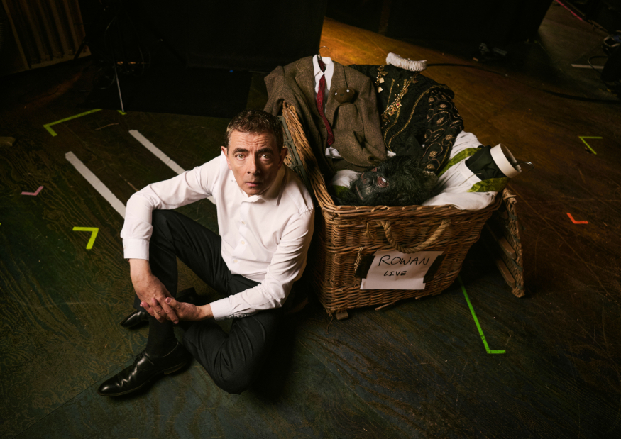 Rowan Atkinson sitting on the floor next to a basket full of stage props