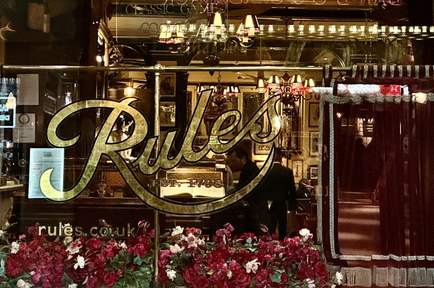 The gold-leaf logo for Rules Restaurant, established in 1798, is displayed on a glass window overlooking a warm, wood-paneled interior with red flowers in the foreground.
