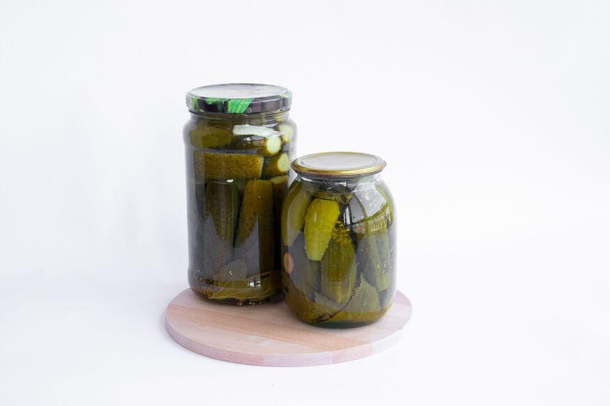Two glass jars of pickled cucumbers on a round wooden board against a white background.