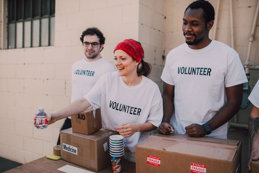 Three smiling volunteers in white t-shirts stand behind a table with boxes of aid, medicine, and water, handing out supplies.